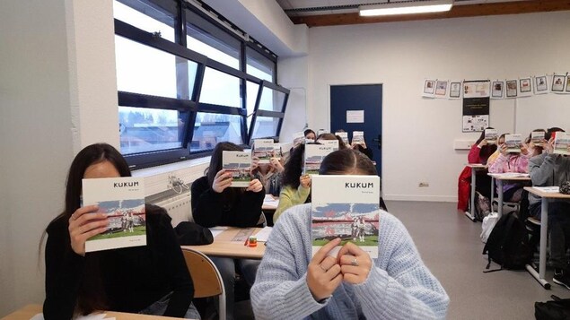 Les jeunes assis et assises à leur pupitre dans une salle de classe montrent le livre de manière à cacher leur visage.