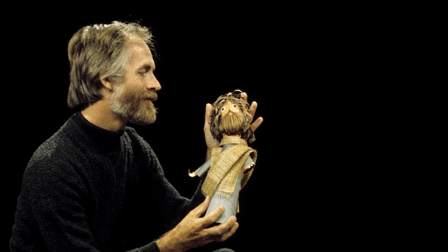 Dans un studio de télévision, Claude Lafortune avec un personnage en papier dans ses mains.