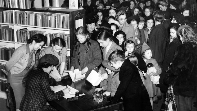 L'autrice est assise à une table et fait des signatures dans des livres. Une foule d'enfants est devant elle, parmi les bibliothèques de livres du magasin. 