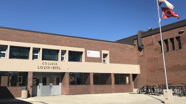 Une école en briques, vue de l'extérieur, un jour de printemps ensoleillé. En avant : les drapeaux franco-manitobain et métis flottent au vent.