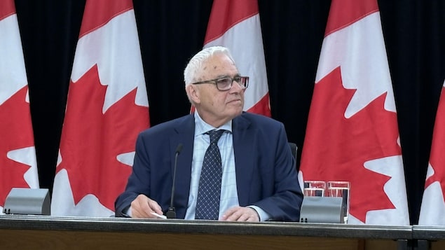 Le commissaire aux langues officielles dans une conférence de presse devant des drapeaux canadiens.
