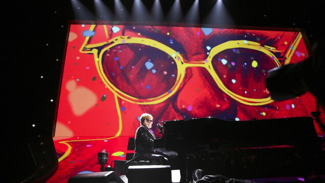 Elton John est au piano. Derrière lui se trouve une grande peinture d'une partie de son visage. Elton John est au piano. Derrière lui se trouve une grande peinture d'une partie de son visage.