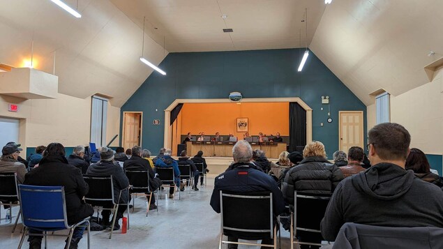 Plusieurs personnes sont assise dans une salle devant une scène où sont installés des conseillers municipaux et un maire.