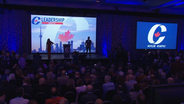 Des personnes sont sur scène devant une projection où l'on peut voir le logo du Parti conservateur du Canada (un « C » entourant une feuille d'érable rouge). Une petite foule les regarde.