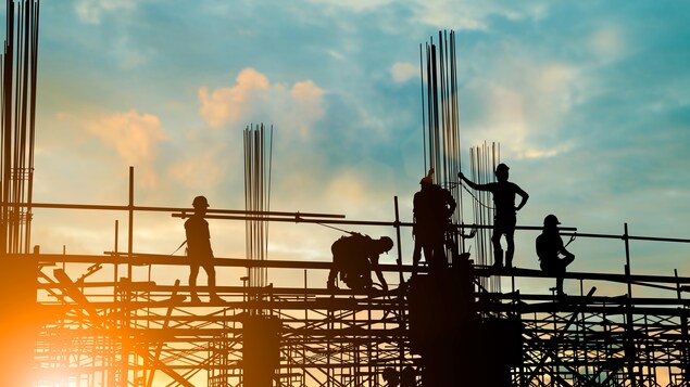 Les silhouettes de personnes sur une partie surélevée d'un chantier de construction sont vues lors d'un coucher ou lever du soleil. Ces personnes portent des casques protecteurs.