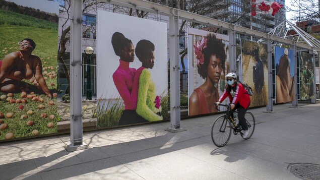 Une femme passe en vélo devant des photographies affichées en extérieur.