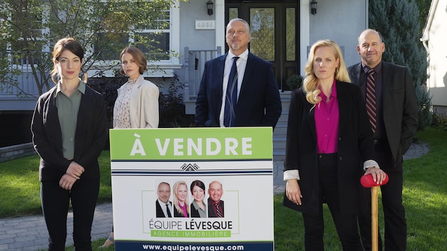 Les cinq personnes se tiennent en rangée, à distance, derrière une affiche de vente immobilière, sur le terrain gazonné devant une maison.