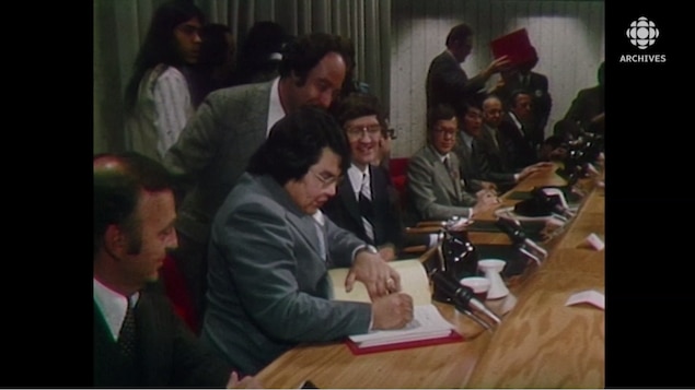Homme qui signe un document sous le regard des autres hommes assis près de lui.