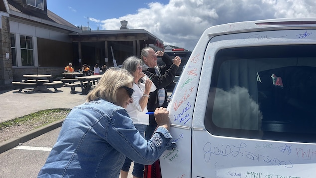 Trois personnes écrivent et signent avec des feutres colorés sur l'arrière d'un véhicule blanc garé à l'extérieur. En arrière-plan, on aperçoit des tables de pique-nique et un bâtiment sous un ciel nuageux.