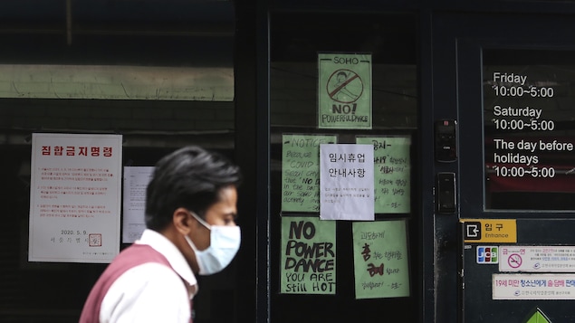 Un homme portant un masque passe devant une devanture avec des affiches signalant la fermeture de l'endroit, une discothèque.