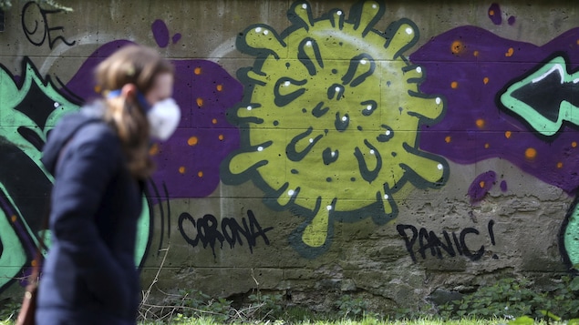 Une femme masquée passe devant un mur de pierre sur lequel est dessiné un graffiti représentant un virus.