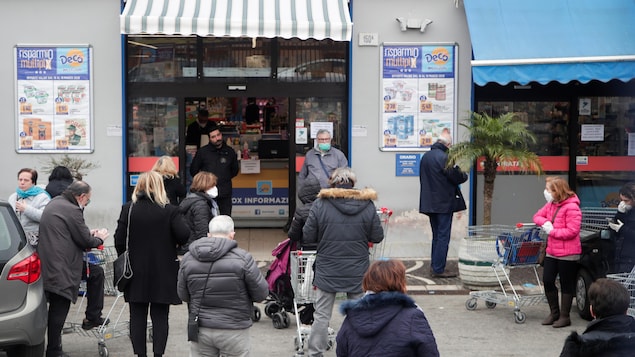 Plusieurs personnes avec des carrosses attendent pour entrer dans un supermarché. Plusieurs sont masquées.