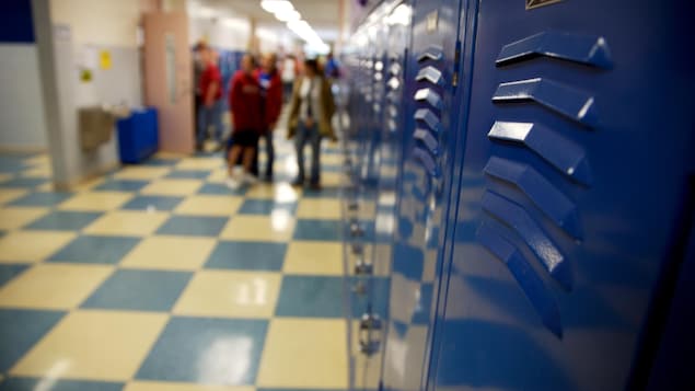 Un corridor d'école. On voit des casiers peints en bleu, un plancher au carrelage jaune et bleu, et des enfants flous au loin.