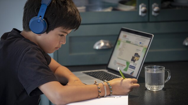 Un élève portant des écouteurs prend des notes sur une feuille devant un ordinateur portable.