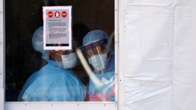 Des infirmières en tenue de protection observent l’extérieur depuis une tente qui a été installée devant un centre hospitalier.