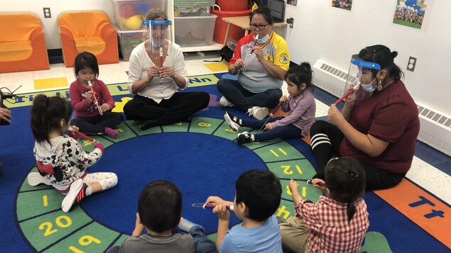 Des enfants et leurs éducatrices sont assis en cercle sur un tapis ludique et jouent de la flûte.