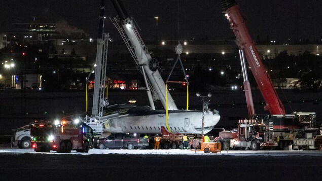 Un avion retourné est soulevé par deux grues pendant la nuit.