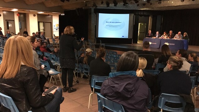 Des parents réunis dans un agora pour une rencontre avec une femme qui pose des questions à un panel.