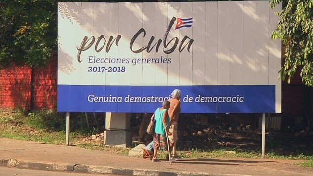 Une grande pancarte, en partie à l'ombre, est vue sur le bord d'une rue. Sur la photographie, deux personnes passent devant elle. Un drapeau de Cuba est par ailleurs dessiné sur la pancarte et les mots sur celle-ci sont en espagnol. Une grande pancarte, en partie à l'ombre, est vue sur le bord d'une rue. Sur la photographie, deux personnes passent devant elle. Un drapeau de Cuba est par ailleurs dessiné sur la pancarte et les mots sur celle-ci sont en espagnol.