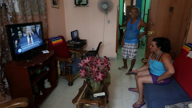 Deux femmes, une assise sur un divan, regarde un écran sur lequel on peut voir Donald Trump parler.