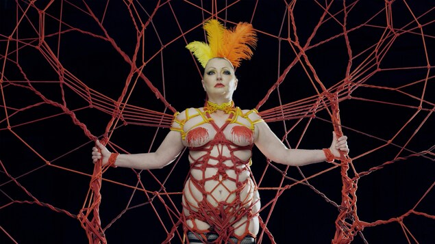 Une femme presque nue se tient dans un décor de mailles rouges, avec un chapeau en plumes.
