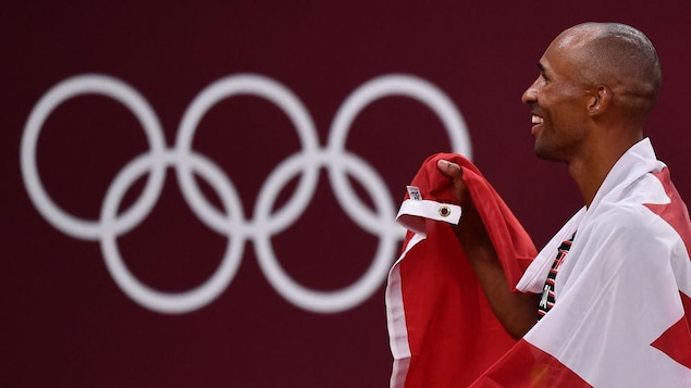 Damian Warner sourit, le drapeau canadien lui couvrant les épaules comme une cape.