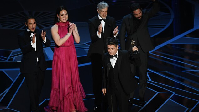 Le réalisateur chilien Sebastián Lelio brandit son trophée sur la scène des Oscars 2018 tandis que quatre membres de son équipe l'applaudissent en arrière. Le réalisateur chilien Sebastián Lelio brandit son trophée sur la scène des Oscars 2018 tandis que quatre membres de son équipe l'applaudissent en arrière.