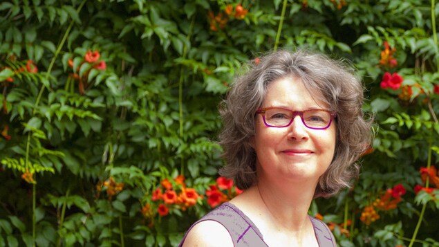 Portrait de la poète Danielle Robitaille avec du feuillage et des fleurs en trompette orange en arrière-plan. Elle a les cheveux gris lâchés et porte des lunettes rouge et rose.