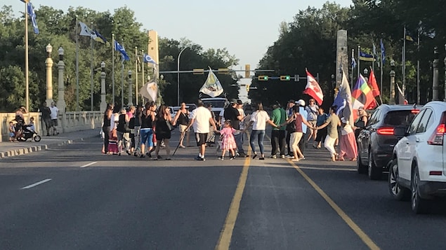 Des gens se tiennent par la main et danse en formant un cercle sur un pont de la ville de Regina, des voitures ne peuvent passer. Des gens se tiennent par la main et danse en formant un cercle sur un pont de la ville de Regina, des voitures ne peuvent passer.