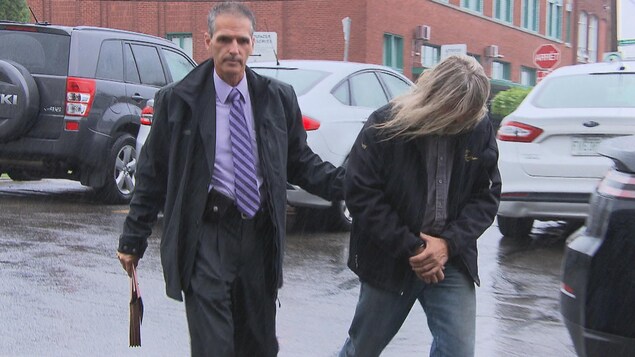 Dany Aubry, la tête baissée, caché par ses cheveux, escorté par un policier dans le stationnement du palais de justice