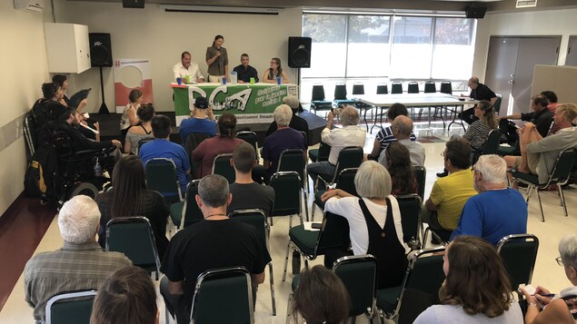Les quatre candidats sont devant une table sur une scène, face à des dizaines de personnes. Isabelle Lévesque est debout et parle au micro. Les quatre candidats sont devant une table sur une scène, face à des dizaines de personnes. Isabelle Lévesque est debout et parle au micro.