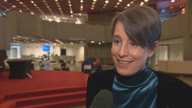 Debi Daviau répond aux questions d'une journaliste dans une grande salle. Debi Daviau répond aux questions d'une journaliste dans une grande salle.