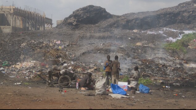 Quatre enfants se tiennent près d'une décharge de déchets au Mali. À gauche de l'image le toit d'une maison est à la même hauteur que le somment de la montagne de déchets. Quatre enfants se tiennent près d'une décharge de déchets au Mali. À gauche de l'image le toit d'une maison est à la même hauteur que le somment de la montagne de déchets.