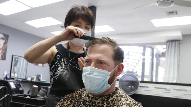 Une coiffeuse peigne les cheveux d'un client. Les deux portent des masques sur le visage.