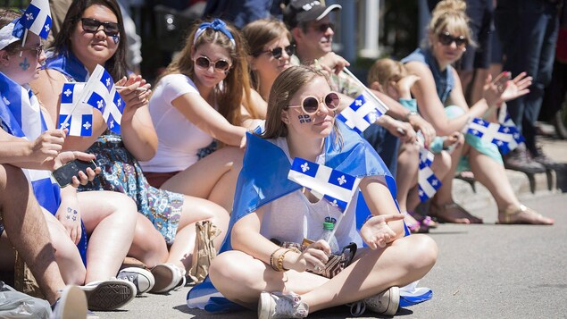 De jeunes filles brandissant des drapeaux du Québec sont assises dans la rue pour assister au défilé de la Fête nationale.