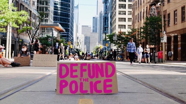 Un panneau « Defund the police » au milieu de la rue Bay, occupée par des manifestants assis.