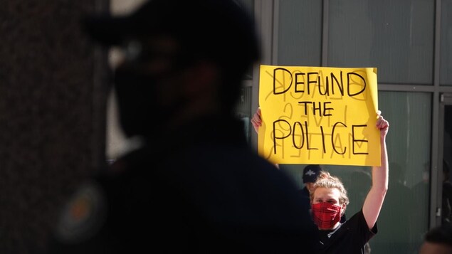 Un homme tient une pancarte sur laquelle est écrit « Defund the police » devant un policier.