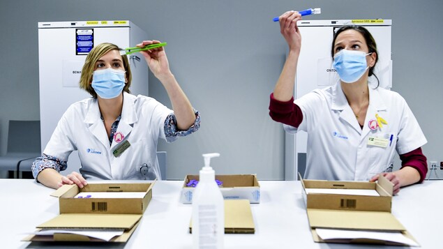 Deux travailleuses de la santé sont assises à une table. Elles lèvent  au-dessus de leurs têtes des fioles de vaccin en utilisant des pinces, et elles en regardent le contenu.