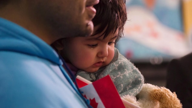 Une petite fille dans les bras de son père qui tient un petit drapeau canadien.