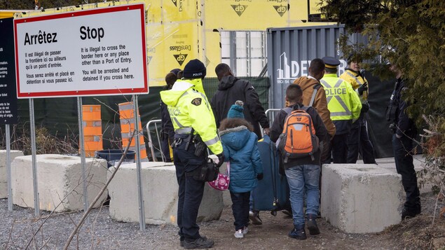Une famille que l'on voit de dos avec sacs et valises est arrêtée par les policiers.