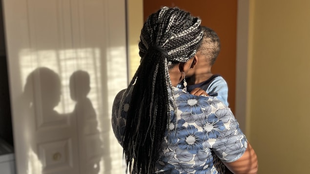 Une femme de dos tient son enfant dans ses bras, leur silhouette se découpe sur la porte.