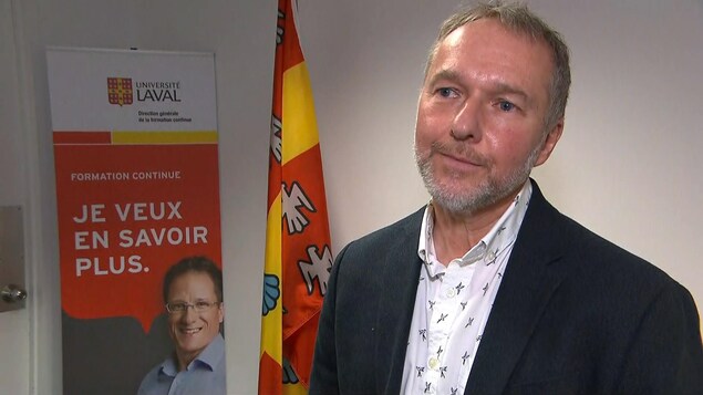 Denis Leclerc avec un complet foncé et une chemise blanche. Il est dans une petite salle avec des bannière de l'Université Laval derrière lui.