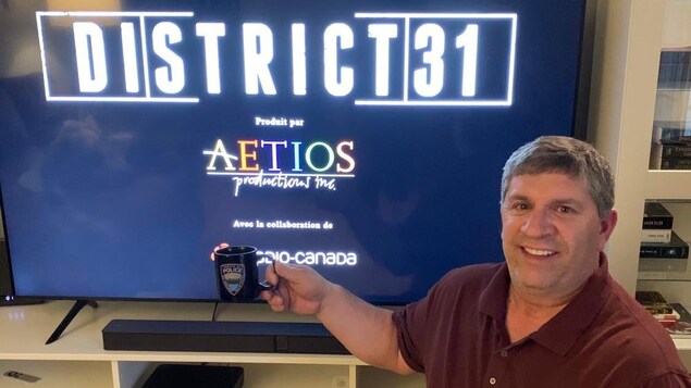 Denis Rioux qui tient sa tasse devant la télé qui affiche District 31