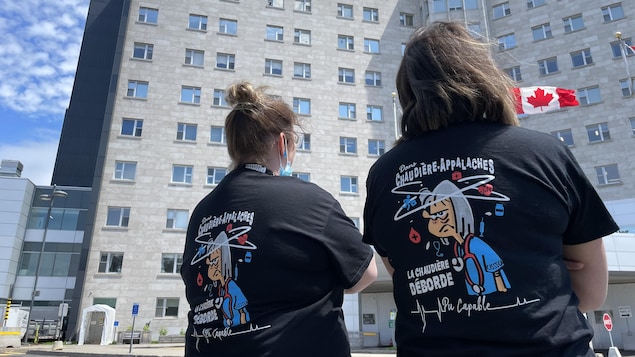 Deux femmes de dos arborant un chandail noir sur lequel est écrit «La Chaudière déborde». En arrière-plan, la façade d'un hôpital.