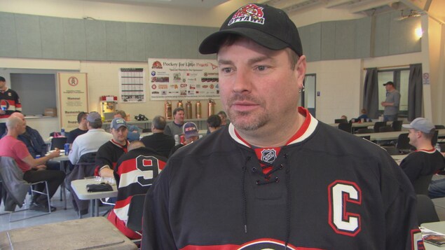 Un homme portant un chandail de hockey dans une salle de réception où sont assises plusieurs personnes. Un homme portant un chandail de hockey dans une salle de réception où sont assises plusieurs personnes.