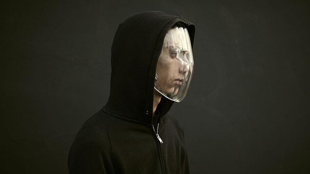 Un homme avec un capuchon noir porte un masque en plastique transparent.