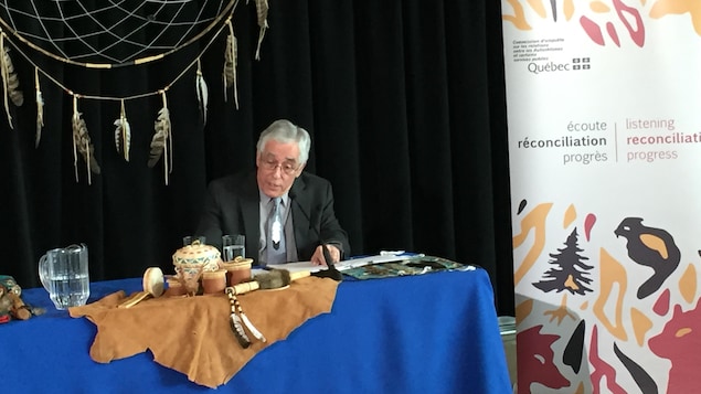 Le commissaire Jacques Viens parle lors d'une conférence de presse, assis à une table où sont déposés des objets traditionnels de communautés autochtones. 