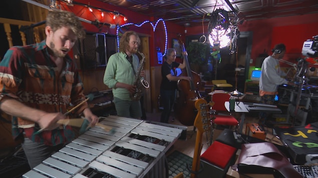 Quatre musiciens sont dans un studio de musique éclectique, aux murs rouges et avec des lumières colorées. Au premier plan, un homme barbu joue du xylophone. Derrière lui, un autre homme joue du saxophone, une femme la contrebasse, et un quatrième homme est à droite, manipulant des platines de DJ.