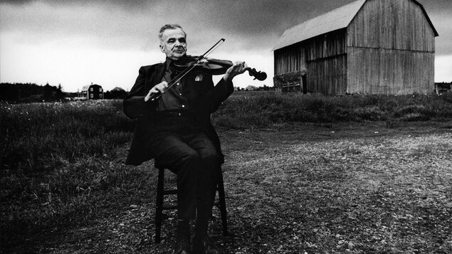 Un homme joue du violon à l'extérieur. On voit une grange en arrière-plan. La photo est en noir et blanc.