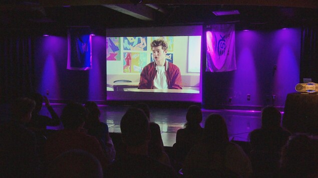 Un écran vidéo devant des jeunes assis au premier plan.